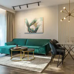 Butelkowa zieleń sofy i dodatki do salonu tworzą elegancką przestrzeń. Szare krzesła i szklany stół dodają nowoczesności.