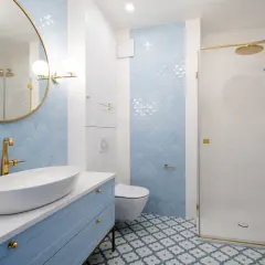 Elegancka łazienka z błękitnymi płytkami w łuski, błękitną szafką i złotą armaturą. Centralnym punktem jest lustro okrągłe złote nad umywalką.