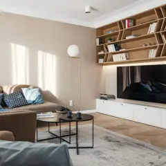 Przytulny salon z sofą i regałem, gdzie dominują kolory pasujące do beżu i brązu. Jasne ściany i drewniane elementy tworzą ciepłą atmosferę.
