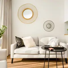 Elegancki salon z jasną kanapą, ozdobiony dwoma okrągłymi lustrami ściennymi. Duże lustro z złotą ramą przyciąga wzrok.
