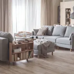 Przytulny salon w beżach i szarościach. Duża, szara sofa z poduszkami, stolik kawowy, regał z książkami i dywanem.