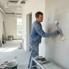 Budowlaniec nakłada gładź na ścianę, sprawdzając, który gips szybciej schnie. W pomieszczeniu widać okna, materiały budowlane i narzędzia.
