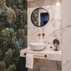Łazienka z tapetą w leśny wzór i marmurowymi płytkami. Złote baterie i lustro tworzą elegancką aranżację.