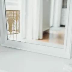 Eleganckie lustro z białą ramą odbija wiklinową lampę i jasne zasłony, tworząc przytulną atmosferę.