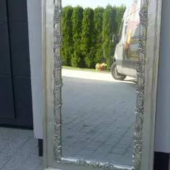 Eleganckie lustro w bogato zdobionej, srebrnej ramie odbija zielone drzewa i zaparkowaną furgonetkę.
