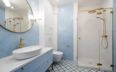 Elegancka łazienka z błękitnymi płytkami w łuski, błękitną szafką i złotą armaturą. Centralnym punktem jest lustro okrągłe złote nad umywalką.