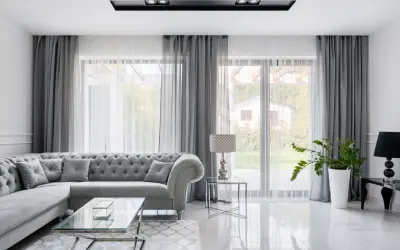 Elegancki salon z szarą sofą i stolikami ze szkła i chromu. W tle okna z zasłonami i lustro glamour srebrne.