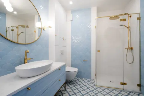 Elegancka łazienka z błękitnymi płytkami w łuski, błękitną szafką i złotą armaturą. Centralnym punktem jest lustro okrągłe złote nad umywalką.