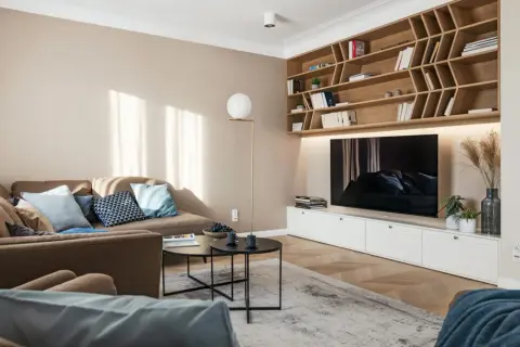 Przytulny salon z sofą i regałem, gdzie dominują kolory pasujące do beżu i brązu. Jasne ściany i drewniane elementy tworzą ciepłą atmosferę.