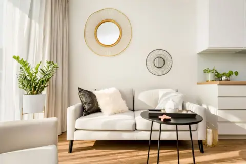 Elegancki salon z jasną kanapą, ozdobiony dwoma okrągłymi lustrami ściennymi. Duże lustro z złotą ramą przyciąga wzrok.