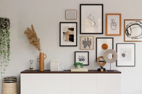 Galeria obrazów nad komodą w salonie, w tym abstrakcje, grafiki i sztuka liniowa. Wśród dekoracji wazon z trawami, klepsydra i wisząca roślina.