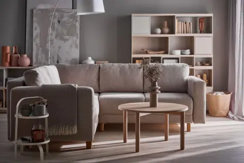 Przytulny salon szaro beżowy inspiracje: wygodna kanapa, okrągły stolik, regał z dekoracjami i kosz z tkanin.