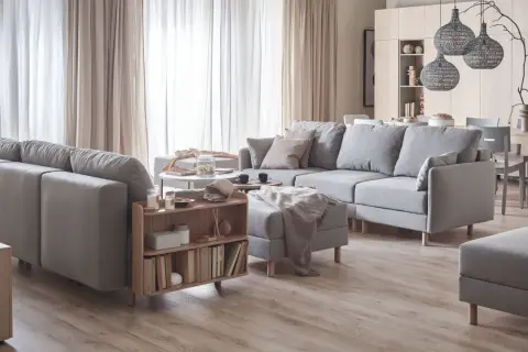 Przytulny salon w beżach i szarościach. Duża, szara sofa z poduszkami, stolik kawowy, regał z książkami i dywanem.