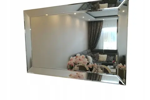 Eleganckie lustro fazowane w ramie, odbijające salon z kanapą i kwiatami.