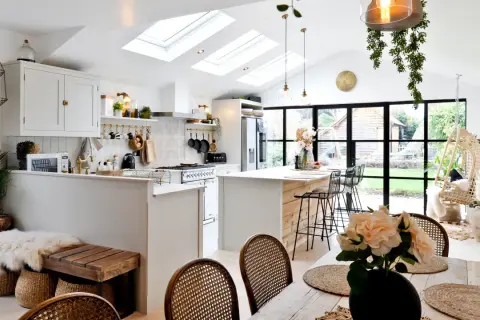 Jasna kuchnia w stylu modern farmhouse z wyspą, białymi meblami i drewnianymi akcentami. Dachowe okna wpuszczają mnóstwo światła.