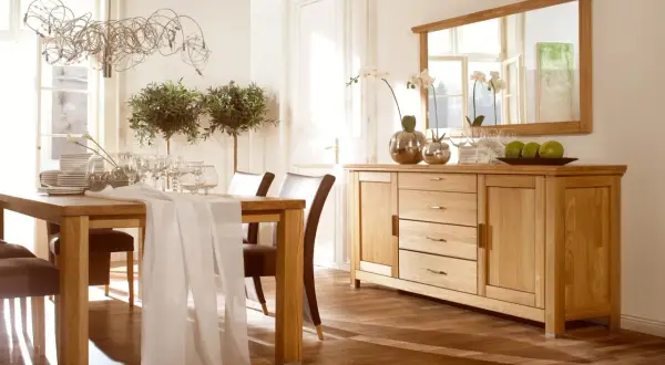 Elegancka jadalnia z drewnianym stołem i krzesłami. Nad komodą wisi duże lustro z drewnianą ramą, odbijające światło i okna.