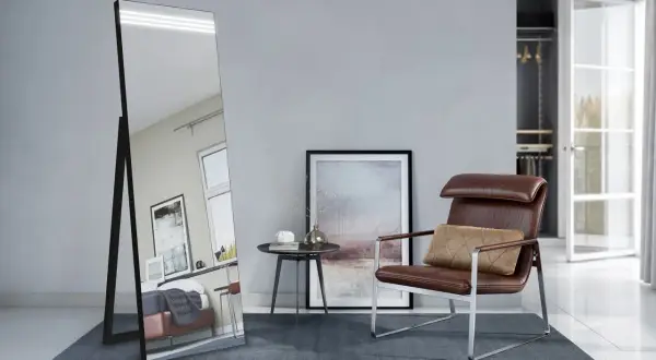 Eleganckie, duże lustro stojące w nowoczesnym wnętrzu, odbijające łóżko i okno. Obok fotel i stolik.
