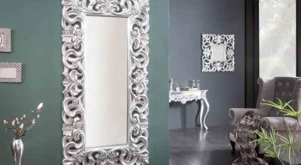 Eleganckie, wysokie lustro w srebrnej ramie z bogatymi zdobieniami, stojące obok wazonu z gałązkami.