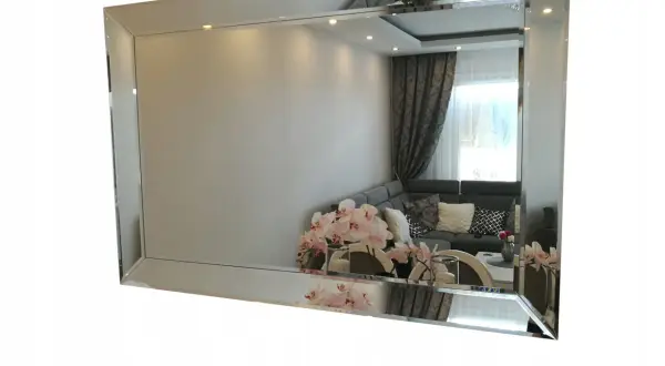 Eleganckie lustro fazowane w ramie, odbijające salon z kanapą i kwiatami.