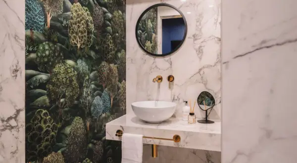 Łazienka z tapetą w leśny wzór i marmurowymi płytkami. Złote baterie i lustro tworzą elegancką aranżację.