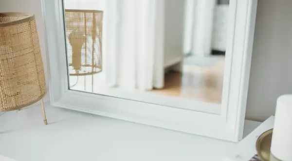 Eleganckie lustro z białą ramą odbija wiklinową lampę i jasne zasłony, tworząc przytulną atmosferę.