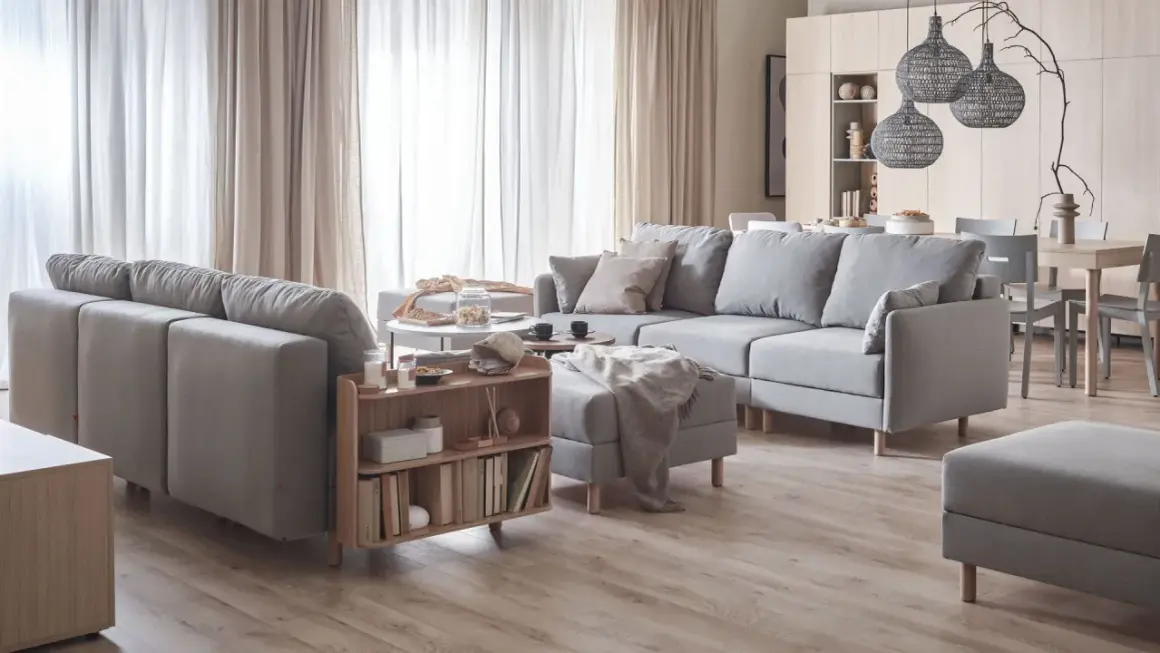 Przytulny salon w beżach i szarościach. Duża, szara sofa z poduszkami, stolik kawowy, regał z książkami i dywanem.