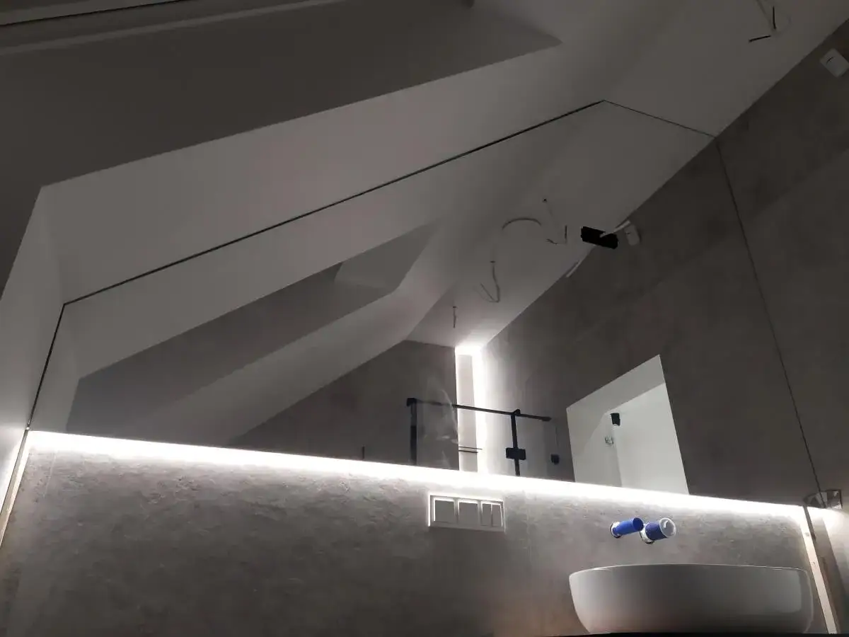 Nowoczesne lustro do łazienki led z wbudowanym oświetleniem, odbijające skośny sufit i umywalkę.