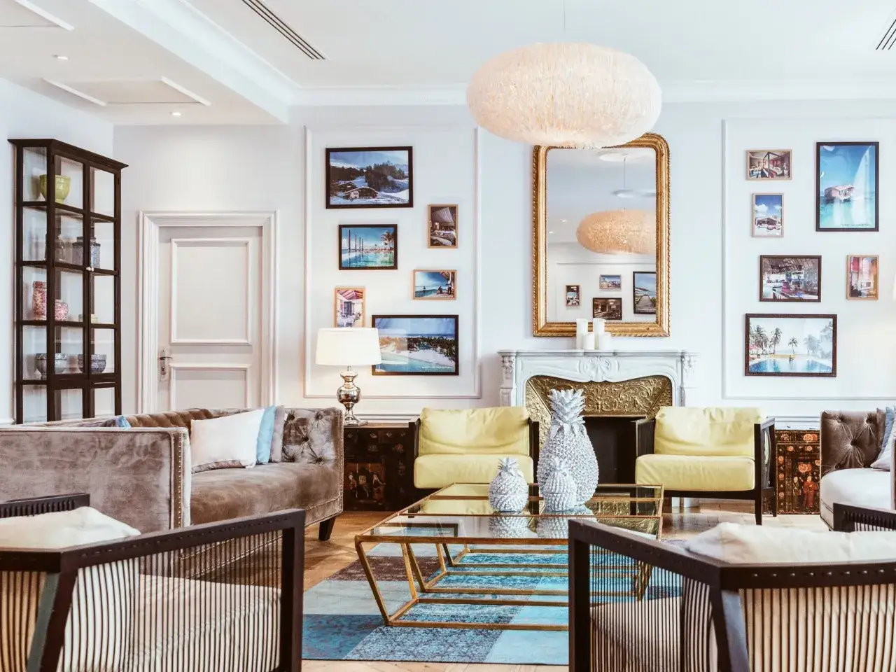 Elegancki salon z sofą, żółtymi fotelami i kominkiem. Nad nim wisi duże lustro ze złotą ramą, odbijające lampę.