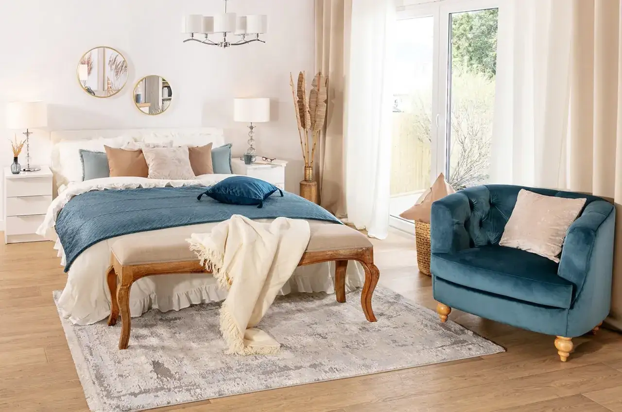 Sypialnia w stylu new hampton: łóżko z niebieską narzutą, ławka, fotel i dywan.