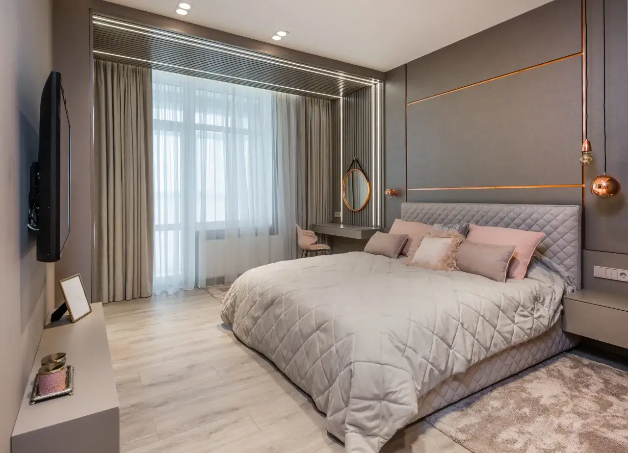 Elegancka sypialnia z łóżkiem i poduszkami w odcieniach brudny róż. Szare ściany z miedzianymi akcentami i nowoczesne oświetlenie tworzą przytulną atmosferę.