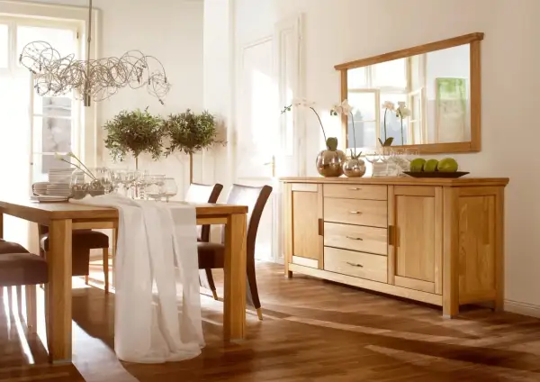 Elegancka jadalnia z drewnianym stołem i krzesłami. Nad komodą wisi duże lustro z drewnianą ramą, odbijające światło i okna.