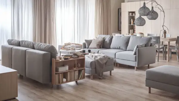 Przytulny salon w beżach i szarościach. Duża, szara sofa z poduszkami, stolik kawowy, regał z książkami i dywanem.