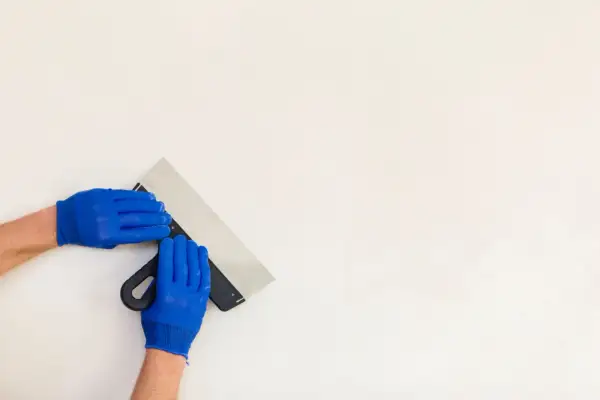 Ręce w niebieskich rękawiczkach trzymają szpachelkę, przygotowując się do pracy. Czas, ile schnie gips ceramiczny, zależy od warunków.